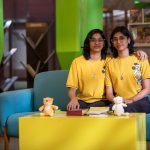 Digital Twins: Redefining Operational Certainty for Executives Twin sisters sitting on a colorful sofa in a library, smiling and wearing yellow t-shirts with cartoon prints.