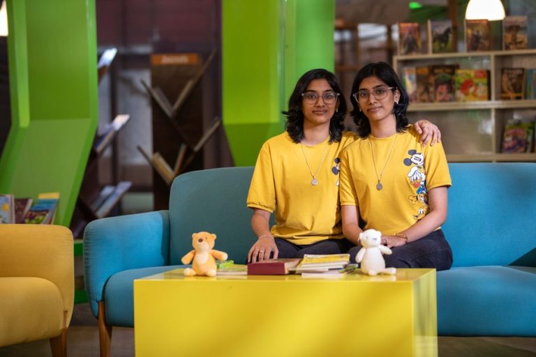 Twin sisters sitting on a colorful sofa in a library, smiling and wearing yellow t-shirts with cartoon prints.