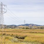 Power lines stretch across a picturesque meadow with hills in the background under a clear sky.