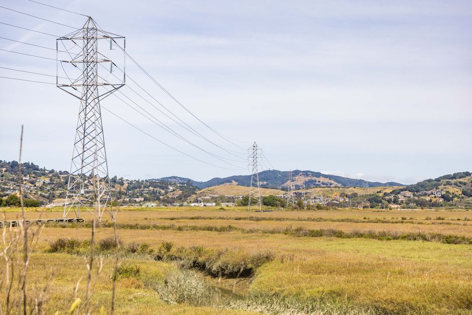 Power lines stretch across a picturesque meadow with hills in the background under a clear sky.
