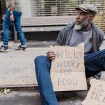 Economic Inequality: A Strategic Constraint on Performance African American man holding a sign seeking help on the sidewalk, representing poverty and homelessness.