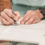 Close-up of a man signing an official document with a ballpoint pen on a desk.