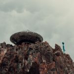 Extraterrestrial Governance: Rethinking Space Law and Strategy A surreal scene with a UFO and alien standing atop a rugged rock formation against a cloudy sky.
