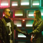 Two individuals with prosthetic arms engage in a handshake amidst a vibrant, retro television backdrop.