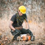 Man wearing safety gear checks chainsaw in autumn forest clearing.