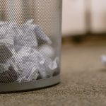 Close-up image of a mesh wire garbage can with scattered crumpled papers on a carpeted floor.