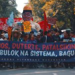 Street protest with demonstrators holding political banners and signs demanding change.