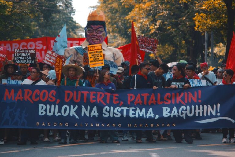 Street protest with demonstrators holding political banners and signs demanding change.