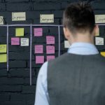A person in a vest stands in front of a project management board with sticky notes on a dark brick wall.