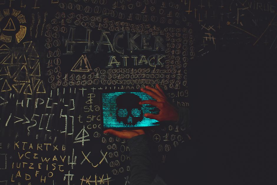 A hacker in a black hoodie using a tablet displaying a skull, surrounded by chalk symbols and 'Hacker Attack' text.