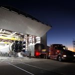Modern cargo truck parked near hangar with massive rocket core inside at industrial factory in evening
