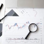 Flat lay of stock market analysis documents with magnifying glass, pens, and glasses.
