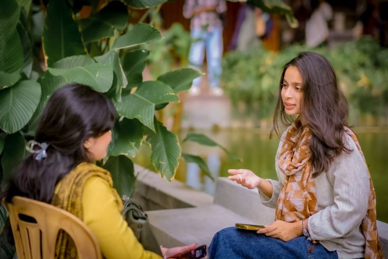 Two women sitting near lush plants having a conversation outdoors. Relaxed and candid atmosphere.