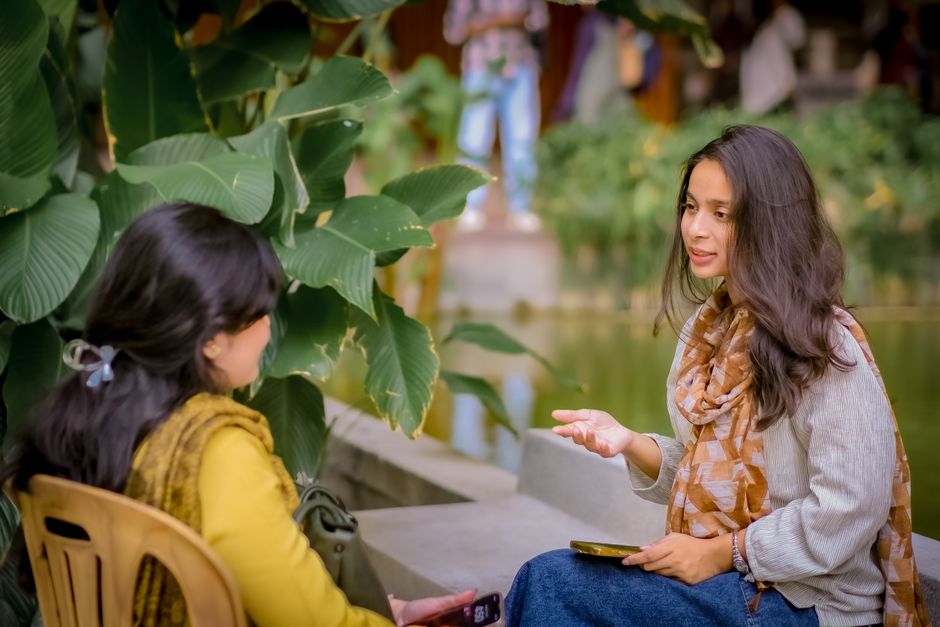 Two women sitting near lush plants having a conversation outdoors. Relaxed and candid atmosphere.