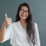 Smiling woman in glasses and white shirt giving a thumbs up against gray background.