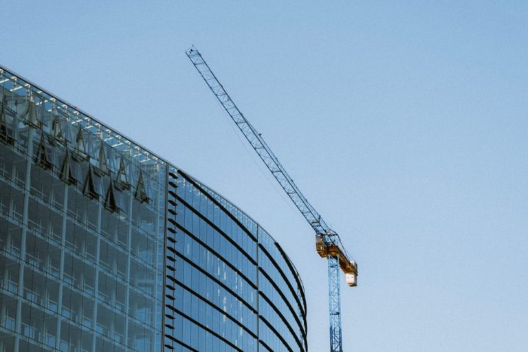 Contemporary glass building under construction with a visible crane against a clear blue sky.