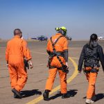 Rescue personnel in orange uniforms walking on airport tarmac with helicopters during the day.