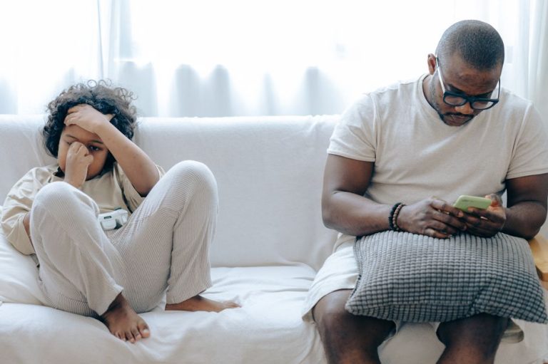 Concentrated African American man in nightwear using smartphone while sitting on cozy sofa with son resting with joystick in light room