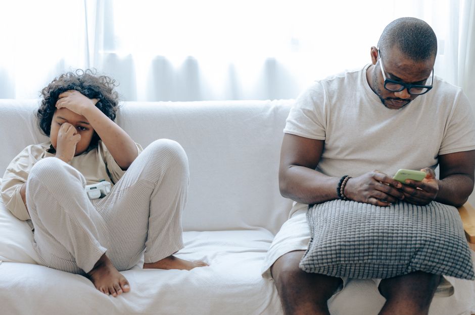 Concentrated African American man in nightwear using smartphone while sitting on cozy sofa with son resting with joystick in light room