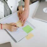 Two people collaborating over a notebook with post-it notes and a laptop in an office setting.