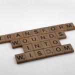 Wooden Scrabble tiles spelling 'Transform Wounds into Wisdom' on a neutral background.
