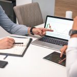 Two businessmen analyzing financial charts and data on a laptop and tablet.