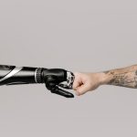 Tactile Robotics: Why Leaders Must Adopt Adaptive Automation Close-up of a tattooed arm fist bumping with a robotic prosthetic arm in studio setting.