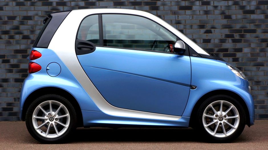 Side view of a modern blue smart car against a brick wall, showcasing compact design.
