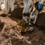 Astronaut planting a small plant on rocky, sandy alien terrain in futuristic space suit.