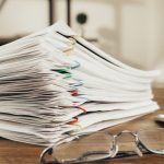 The Administrative Trap: Why Mandates Erode Operational Success Close-up of a stack of office papers on a desk with glasses, emphasizing organization.