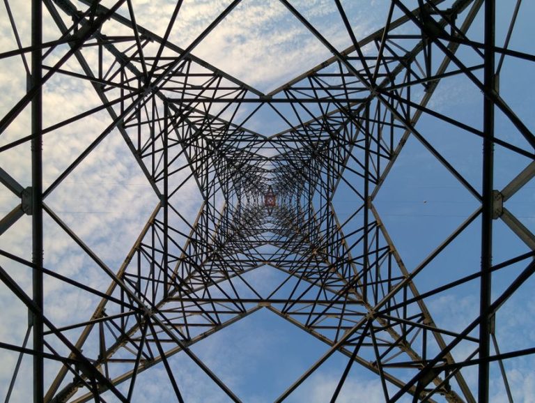 Looking up through a steel power tower against a blue sky and clouds.