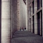 A modern architectural colonnade with tall stone columns and people walking through the space.