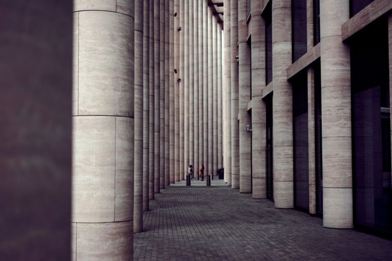 A modern architectural colonnade with tall stone columns and people walking through the space.