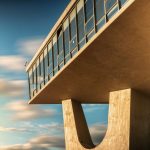 Futuristic building facade with large windows and concrete pillars under a vibrant cloudy sky.