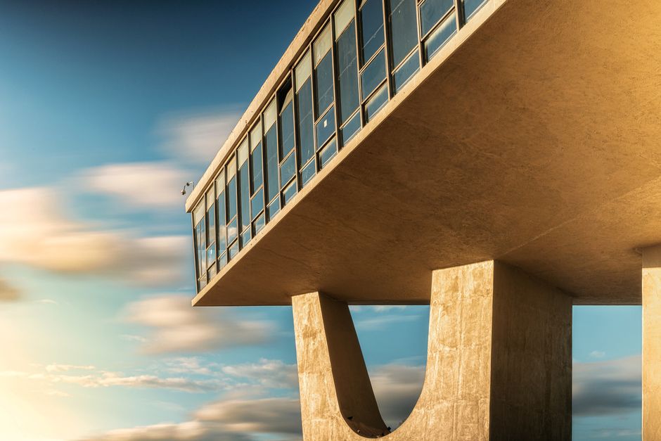 Futuristic building facade with large windows and concrete pillars under a vibrant cloudy sky.