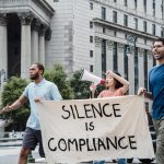The Compliance Trap: Why Political Alignment Can Kill Innovation Activists marching with a banner saying 'Silence is Compliance' outside a courthouse.