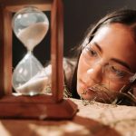 A woman wearing glasses observes an hourglass, symbolizing time and reflection.