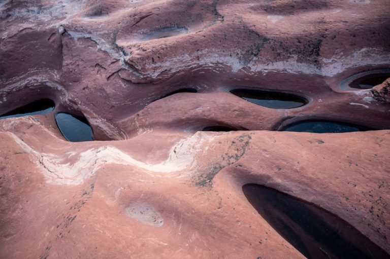 Aerial view of red sandstone formations with unique eroded patterns and water pools.