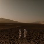 Two astronauts in spacesuits walk across a Mars-like desert landscape during the day.