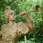 A bronze statue of a warrior playing a trumpet stands amidst lush green trees.