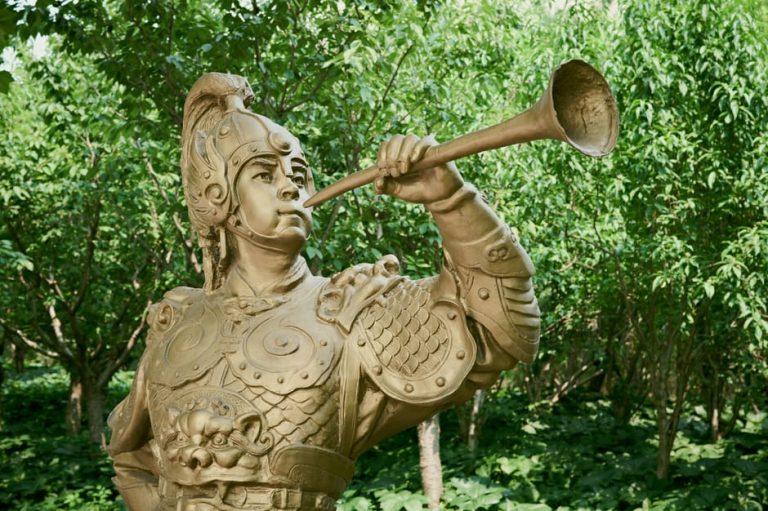 A bronze statue of a warrior playing a trumpet stands amidst lush green trees.