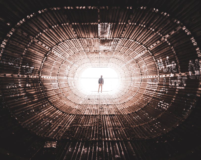 Silhouette of a person standing at the end of a geometric, metallic tunnel with dramatic perspective and lighting.