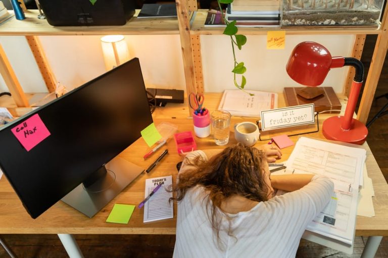 A tired woman rests at her office desk surrounded by work documents and a computer monitor.