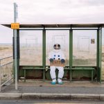 An astronaut in a spacesuit sits at a rural bus stop holding a sign reading 'Earth.'