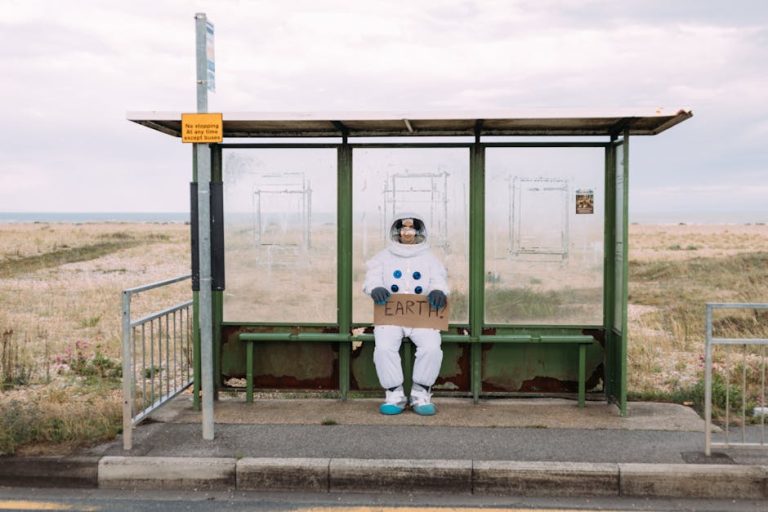 An astronaut in a spacesuit sits at a rural bus stop holding a sign reading 'Earth.'