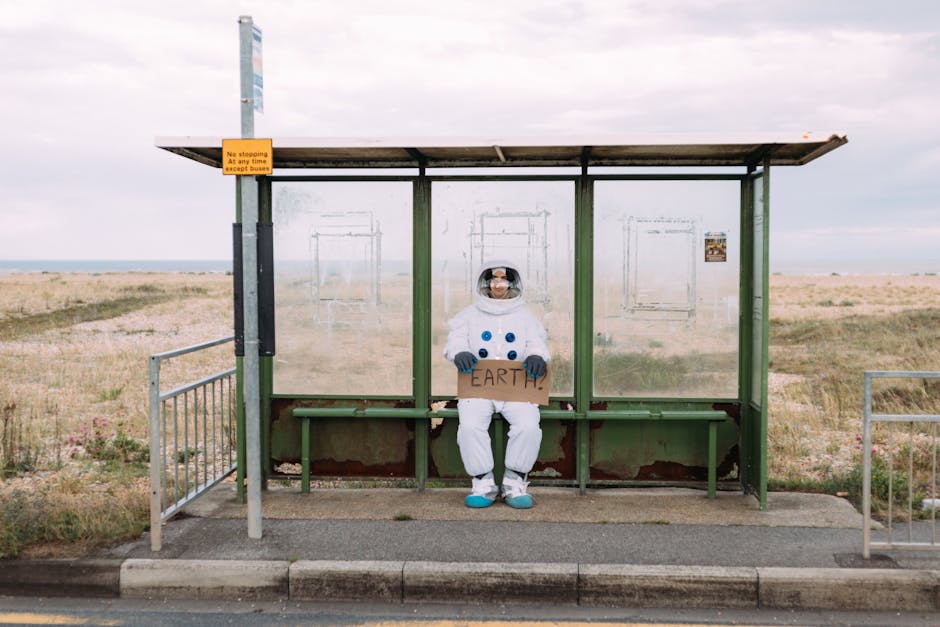 An astronaut in a spacesuit sits at a rural bus stop holding a sign reading 'Earth.'