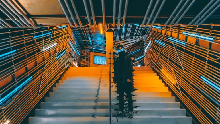 A person stands on a futuristic staircase surrounded by neon lights and metallic walls.