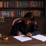 A judge in robes writing on a document at a desk in an office library with law books.