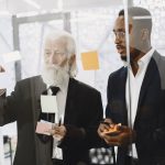 The Mentor’s Paradox: Why Scaling Yourself is the Ultimate Leadership Test Two men in suits discussing ideas with sticky notes on a glass wall in an office.
