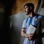 <title>The Messi Protocol: Can Strategic Longevity Sustain a 2026 World Cup Run?</title> Man in Argentina soccer jersey holding book indoors, looking thoughtful.
